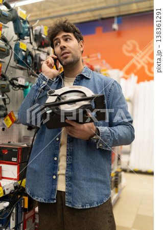 Man talking on phone while holding a circular saw in a tools shop Man talking on phone while holding a circular saw in a tools shop 138361291