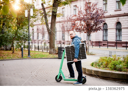 Man rides electric scooter while carrying solar panel. Integration of solar power as sustainable energy source for charging electric scooters, promoting eco-friendly urban transportation. Man rides electric scooter while carrying solar panel. Integration of solar power as sustainable energy source for charging electric scooters, promoting eco-friendly urban transportation. 138362975
