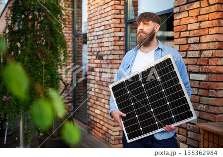 Portrait of bearded man holds solar panel, standing in front of brick building and lush greenery. Guy in shirt and t-shirt exudes calm and eco-conscious vibe, blending nature with urban elements. Portrait of bearded man holds solar panel, standing in front of brick building and lush greenery. Guy in shirt and t-shirt exudes calm and eco-conscious vibe, blending nature with urban elements. 138362984
