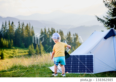 Young child playing near tourist tent with photovoltaic solar panel in summer. On background of mountains and trees under clear blue sky. Integration of renewable energy in outdoor camping activities. Young child playing near tourist tent with photovoltaic solar panel in summer. On background of mountains and trees under clear blue sky. Integration of renewable energy in outdoor camping activities. 138362985