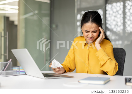 Young business woman feeling intense headache or migraine, holding her temple and a tissue, sitting at a laptop in a modern office setup, experiencing work-related stress, burnout, or illness Young business woman feeling intense headache or migraine, holding her temple and a tissue, sitting at a laptop in a modern office setup, experiencing work-related stress, burnout, or illness 138363226