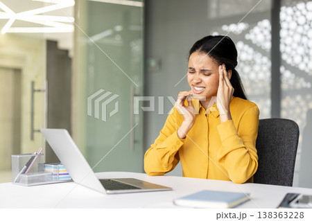 Young woman working on laptop in a modern office, feeling severe headache and pain, touching her temples with a pained expression due to stress and burnout Young woman working on laptop in a modern office, feeling severe headache and pain, touching her temples with a pained expression due to stress and burnout 138363228