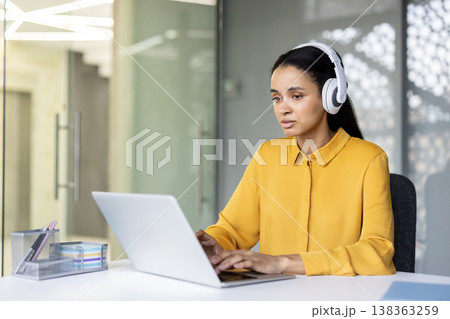 Focused woman wearing white headphones and a yellow shirt, sitting at a desk and typing on a silver laptop, engaging in remote work or online communication in a contemporary office environment 138363259