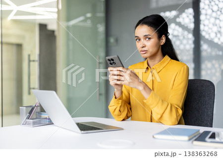 Young professional woman using a smartphone in a modern, bright office with a laptop on the desk, focusing on digital communication and multitasking during her workday 138363268