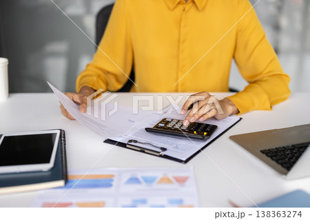 Hands of a person wearing a yellow shirt using a calculator to perform financial calculations while reviewing documents, and preparing taxes on a modern office desk with a laptop and tablet 138363274