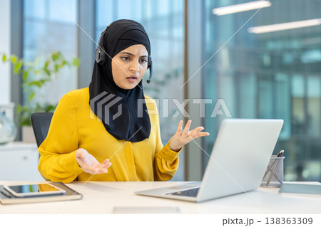 Muslim businesswoman wearing a headset and hijab experiencing frustration and stress during a virtual meeting or customer support call, using a laptop in a modern office environment 138363309