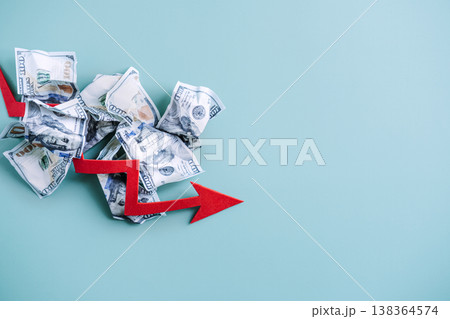 Red arrow points downward over pile of crumpled dollar bills on blue background. Negative economic growth, global financial crisis, bankruptcy warning. 138364574