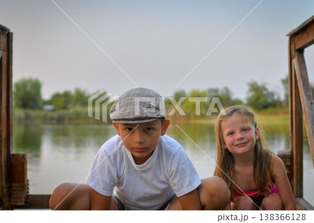 A little boy and sweet girl kneel on a wooden pier on the shore of a lake. Small children sit on a pier at the edge of a pond. The concept of a carefree childhood and summer vacations. 138366128
