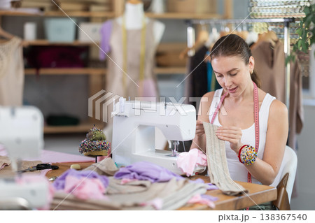 Glad young dressmaker viewing dress detail sitting at sewing-machine 138367540