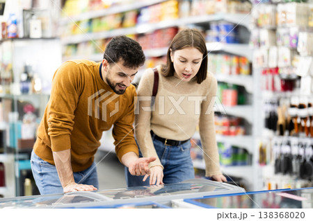 Positive young couple choosing cooled food product standing at fridge in grocery store Positive young couple choosing cooled food product standing at fridge in grocery store 138368020