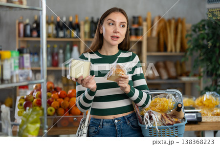Smiling young girl with shopping basket trying to make choice holding two sorts of cheese in grocery store Smiling young girl with shopping basket trying to make choice holding two sorts of cheese in grocery store 138368223