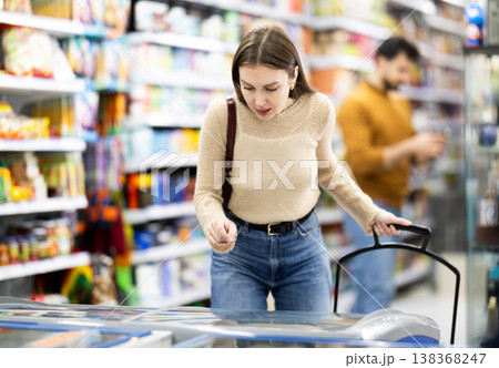 Woman standing near refrigerated display cases choosing frozen food in supermarket 138368247