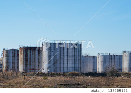 Row of large industrial storage tanks with rust and weathering in open field under blue sky Row of large industrial storage tanks with rust and weathering in open field under blue sky 138369131