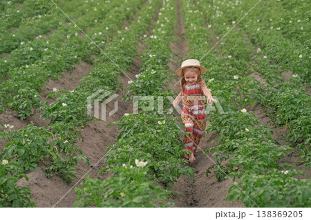 A funny, happy barefoot girl in a hat walks between rows of flowering potatoes. Cultivation of agricultural crops 138369205