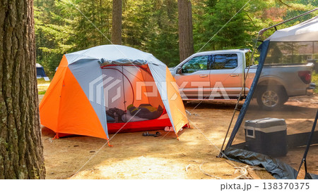 Colorful camping tent set up in a forest clearing with a pickup truck parked nearby and additional camping gear visible in the background Colorful camping tent set up in a forest clearing with a pickup truck parked nearby and additional camping gear visible in the background 138370375