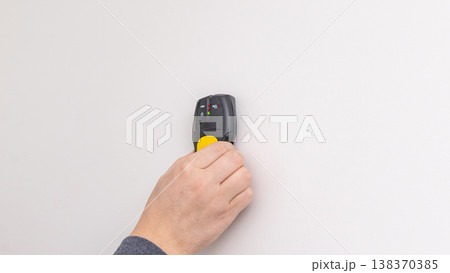 Mans hand pressing yellow and black stud finder onto white wall close to power outlet. Home improvement, detecting studs, DIY process, tool application. Mans hand pressing yellow and black stud finder onto white wall close to power outlet. Home improvement, detecting studs, DIY process, tool application. 138370385