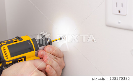 Mans hands using yellow cordless drill with light on, illuminating wall area near outlet during work. Home improvement, drilling with flashlight, DIY task, illuminated workspace. Mans hands using yellow cordless drill with light on, illuminating wall area near outlet during work. Home improvement, drilling with flashlight, DIY task, illuminated workspace. 138370386