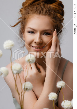 Young Caucasian redhead woman touching face among dried white flowers on moss, white wall Young Caucasian redhead woman touching face among dried white flowers on moss, white wall 138371393