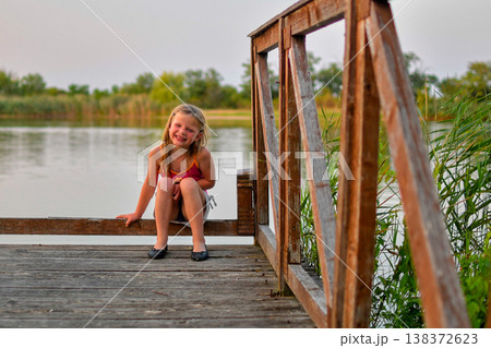 Joyful young girl sitting on a wooden dock by the lake on a summer day 138372623