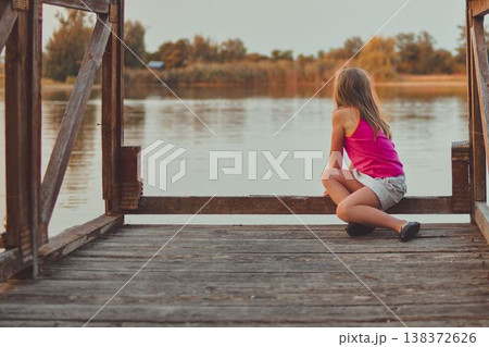 A cute young girl sits on a wooden pier at the end of a lake. The photo was taken during the golden hour, at sunset. It represents childhood, summer vacations, looking toward the future, and carefree A cute young girl sits on a wooden pier at the end of a lake. The photo was taken during the golden hour, at sunset. It represents childhood, summer vacations, looking toward the future, and carefree 138372626