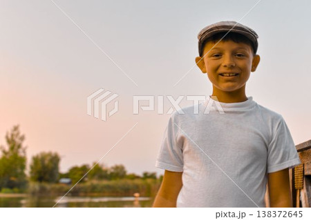 A smiling young boy stands on a pier at the edge of a pond. The concept of summer vacation, carefree days, and childhood. A photo taken at sundown. A smiling young boy stands on a pier at the edge of a pond. The concept of summer vacation, carefree days, and childhood. A photo taken at sundown. 138372645