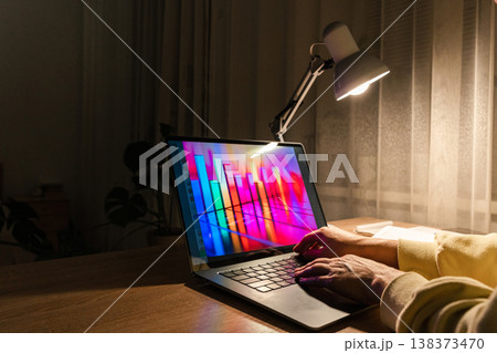 Hands typing on laptop keyboard with colorful bar chart displaying on screen, illuminated by desk lamp at night. Person working at night with laptop showing data chart Hands typing on laptop keyboard with colorful bar chart displaying on screen, illuminated by desk lamp at night. Person working at night with laptop showing data chart 138373470