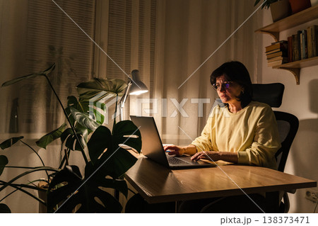 Woman focusing on work while sitting desk with laptop lamp illuminating her home office workspace, working on laptop late night 138373471
