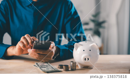 Man uses digital savings mobile payment bank app phone online to save, white piggy bank sits beside coins and cash for payment financial planning future smartphone pig growth Man uses digital savings mobile payment bank app phone online to save, white piggy bank sits beside coins and cash for payment financial planning future smartphone pig growth 138375818