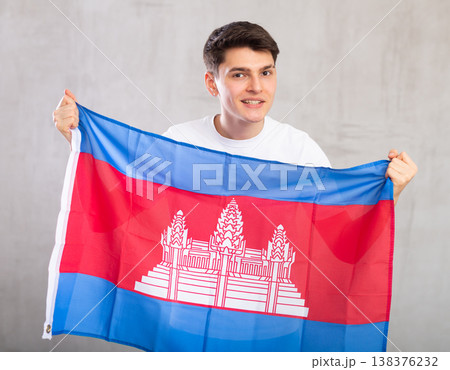 Man in casual clothes holds unfurled flag of Cambodia in hands against gray wall Man in casual clothes holds unfurled flag of Cambodia in hands against gray wall 138376232