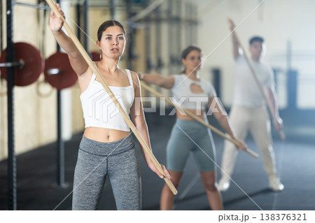 Slender girl, together with other athletes, does a warm-up using a long stick in gym Slender girl, together with other athletes, does a warm-up using a long stick in gym 138376321