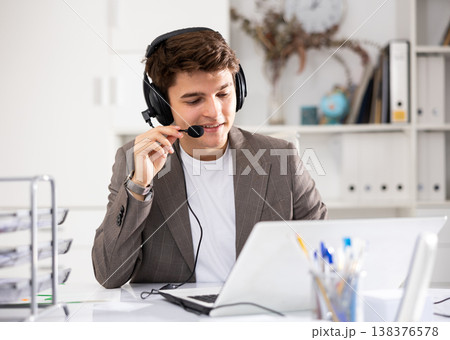 Positive businessman with headphones and microphone having video call on laptop in office 138376578
