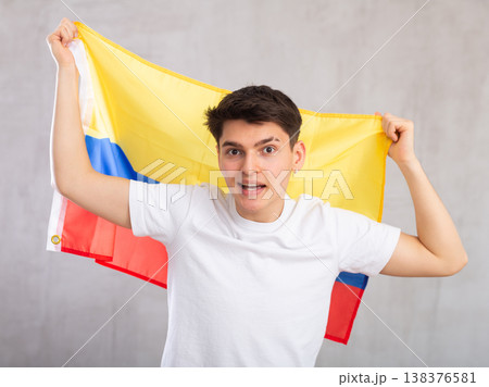 Man in holds unfurled flag of Colombia in hands raised above head against gray wall 138376581