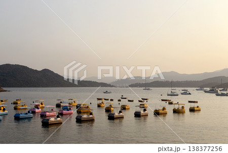 Colorful paddle boats and sailboats dot a calm bay at sunset with rolling hills 138377256