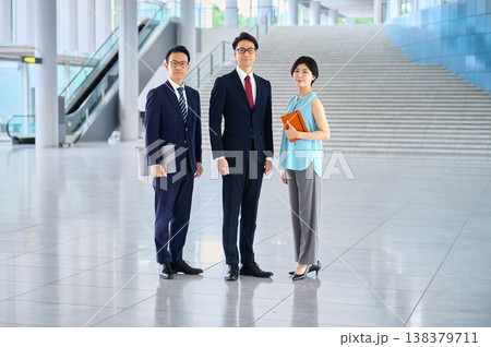 オフィスビルの広々としたロビーに立つ、笑顔で自信に満ちた3人の日本人ビジネスチーム 138379711