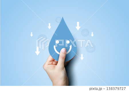 Hand holding a smiling blue water drop icon with downward arrows on blue background. Concept of reducing water consumption, saving water, environmental conservation, and eco-friendly sustainability. 138379727