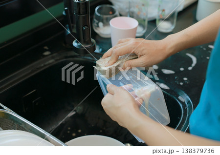 Close up view of hands washing a plastic food storage container with a soapy sponge at the kitchen sink. Woman doing household chores, cleaning dirty dishes in soapy water. Daily domestic routine. Close up view of hands washing a plastic food storage container with a soapy sponge at the kitchen sink. Woman doing household chores, cleaning dirty dishes in soapy water. Daily domestic routine. 138379736