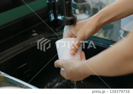 Close up of hands rinsing a white coffee mug under running water from a black faucet at the kitchen sink. Woman washing dishes and doing household chores. Daily domestic routine and cleanliness. Close up of hands rinsing a white coffee mug under running water from a black faucet at the kitchen sink. Woman washing dishes and doing household chores. Daily domestic routine and cleanliness. 138379738