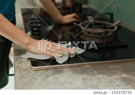 Close up of person hands cleaning a kitchen gas stove with a detergent spray bottle and microfiber cloth. Doing daily household chores, domestic hygiene routine, wiping dirty cooktop surface clean. 138379739