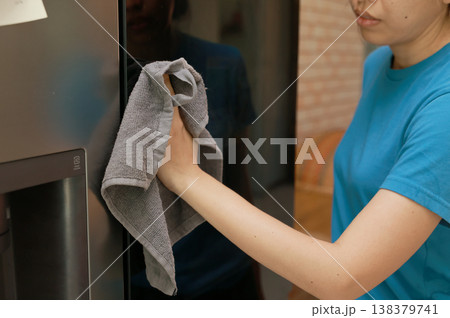 Close up of a woman hand wiping and cleaning a modern refrigerator door with a gray microfiber cloth. Doing daily household chores, domestic hygiene routine, and keeping home kitchen appliances clean. Close up of a woman hand wiping and cleaning a modern refrigerator door with a gray microfiber cloth. Doing daily household chores, domestic hygiene routine, and keeping home kitchen appliances clean. 138379741