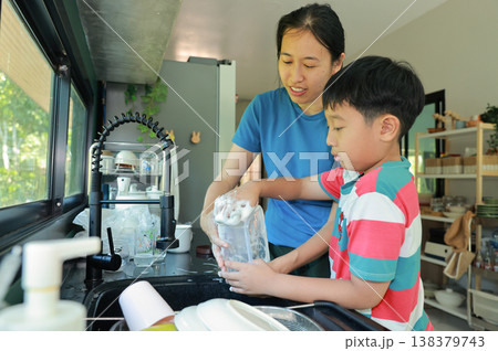 Asian mother teaching her little son how to wash dishes at the kitchen sink. Happy family washing a plastic container with soapy sponge together. Domestic housework, daily chores and parenting. Asian mother teaching her little son how to wash dishes at the kitchen sink. Happy family washing a plastic container with soapy sponge together. Domestic housework, daily chores and parenting. 138379743
