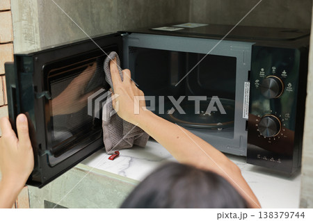 Close up of woman hands wiping and cleaning inside the glass door of a modern black microwave oven with a grey microfiber cloth. Doing daily household chores and keeping kitchen appliances clean. Close up of woman hands wiping and cleaning inside the glass door of a modern black microwave oven with a grey microfiber cloth. Doing daily household chores and keeping kitchen appliances clean. 138379744