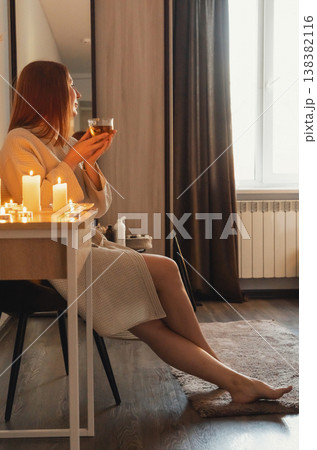 Woman in bathrobe enjoying hot tea illuminated by flickering candles, finding peace and tranquility. Wellness and spa evening Woman in bathrobe enjoying hot tea illuminated by flickering candles, finding peace and tranquility. Wellness and spa evening 138382116