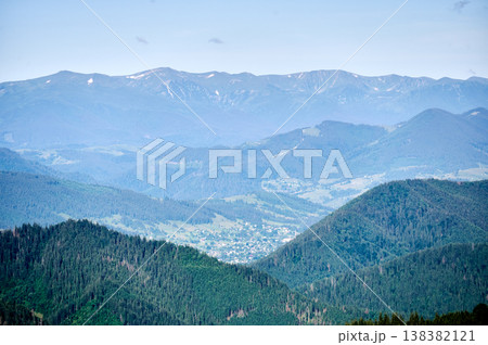 Stunning view of Carpathian Mountains with majestic peaks and deep valleys. Dense forests blanket landscape, leading down to quaint village nestled in valley below. 138382121