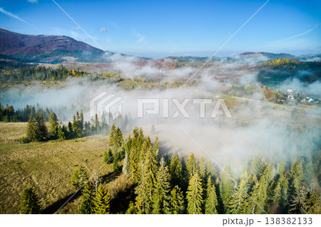 Picturesque view of hillside adorned with vibrant autumn foliage, ranging from rich greens to fiery reds and golden yellows. Wisps of fog drift across landscape under bright blue sky. Aerial view. 138382133