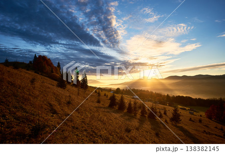 Sun rises over mountains, casting golden rays across landscape. Sky filled with dramatic clouds, creating stunning contrast with clear blue above. Warm light illuminates rolling hills and pine trees. Sun rises over mountains, casting golden rays across landscape. Sky filled with dramatic clouds, creating stunning contrast with clear blue above. Warm light illuminates rolling hills and pine trees. 138382145
