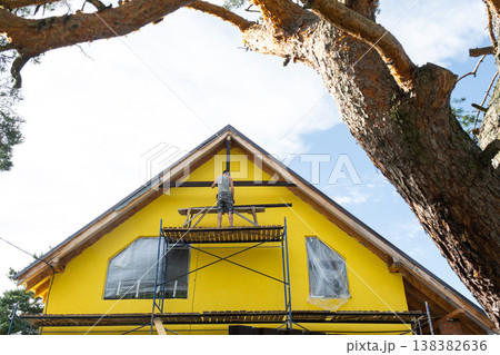 Wide view of bright yellow home with scaffolding and gable work in progress, sunny construction scene, home improvement ongoing. 138382636