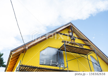 Yellow modern house with gable trim and scaffold network set up, exterior woodwork in process, sunny bright day. 138382638