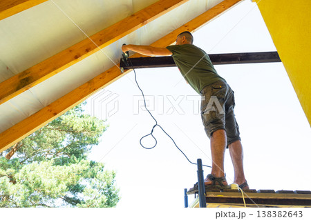 Man with cord fixes black wooden beam inside gable, close-up on hands and timber, modern yellow wall, self-build tasks. Man with cord fixes black wooden beam inside gable, close-up on hands and timber, modern yellow wall, self-build tasks. 138382643