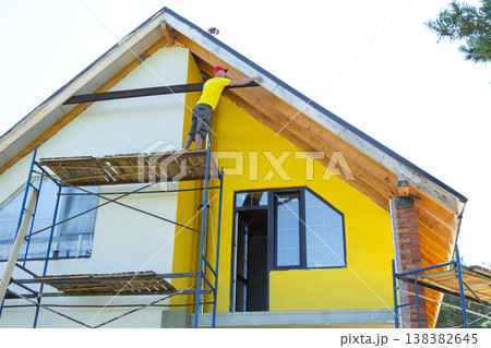 Elevated perspective of worker adjusting upright timber under eave, multi-story yellow house, home upgrade project continues. 138382645