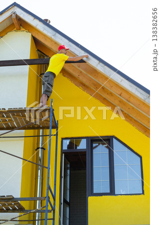 Worker balances on upper scaffold attaching horizontal plank under gable, yellow contemporary facade, critical construction moment, personal safety gear. Worker balances on upper scaffold attaching horizontal plank under gable, yellow contemporary facade, critical construction moment, personal safety gear. 138382656